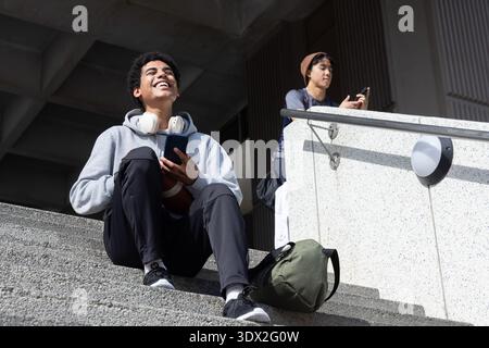 Diverse male friends sitting on campus concrete steps using smartphones and holding football Stock Photo