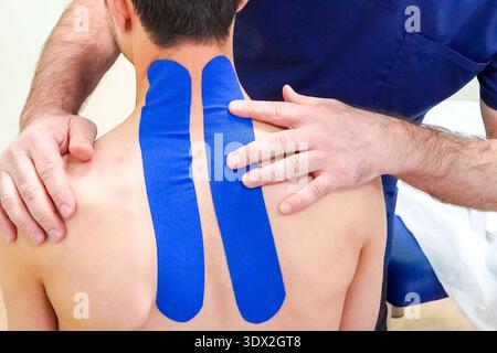 Physiotherapist treats patient neck, back pain with kinesio tape close up. Unrecognizable young caucasian patient.Doctor examining her patient back in Stock Photo