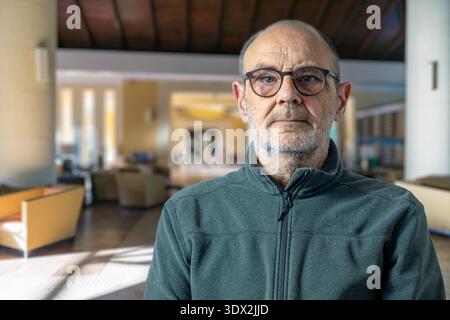 A senior man with round glasses stands in a bright, modern lobby. Calm and thoughtful, he embodies a relaxed, professional mood suitable for business, Stock Photo