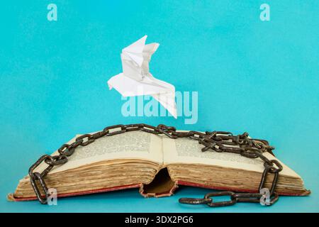 White origami dove bird flying from a opened vintage text book, with broken metal chains on blue background , freedom of speech concept Stock Photo