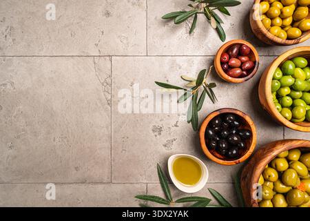 Green and black olives in a bowls and olive oil on gray background top view. Olive oil and variety of green and black olives in bowls with olive leaves all around taken from above. Stock Photo