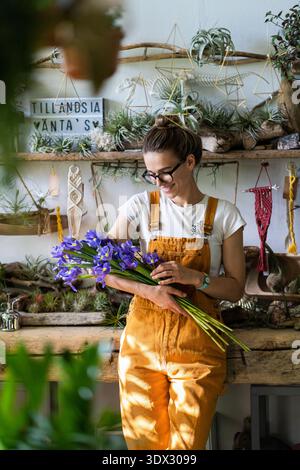 Surprised florist woman in glasses, wear orange overalls surrounded by collection of tillandsia air plants, charmed with pleasant gift, smiling, looking at a bouquet of irises flowers. Indoor garden || Model released Stock Photo