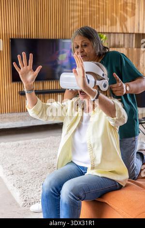 Diverse senior female friends trying out virtual reality headset on orange bench in living room. Technology, innovation, friendship, relaxation, family, modern, leisure || Model released Stock Photo