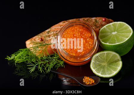 Red salmon trout caviar in glass jar and in spoon with fresh fish steak, lime and dill over glass black background. Stock Photo