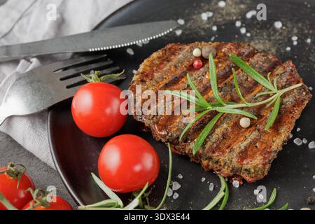 Black Angus Eye of round steak marinated and grilled closeup on black retro plate with rosemary, spices and cherry tomatoes. Spicy tasty beef steak on Stock Photo