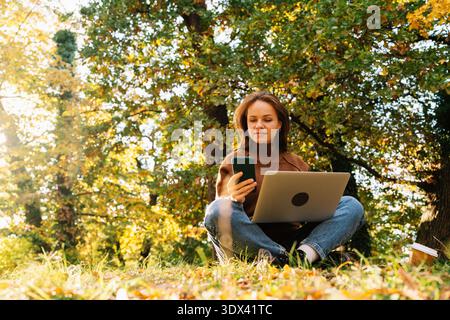 Young adult woman working outdoors in autumn park using laptop and smartphone while sitting on ground. Stock Photo