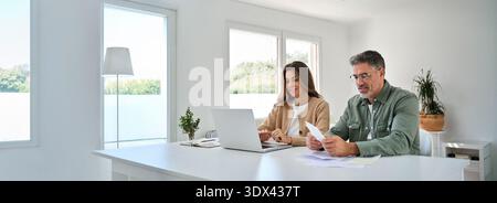 Middle aged mature couple using laptop counting taxes with receipts at home. Stock Photo
