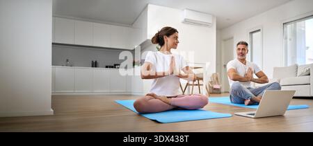 Calm middle aged couple learning online yoga meditating using laptop at home. Stock Photo