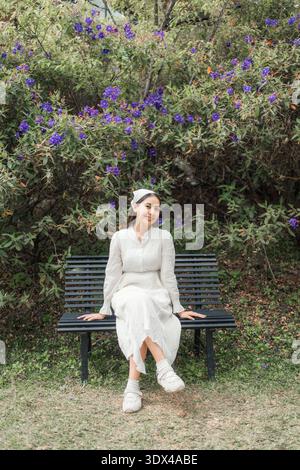 Beautiful asian woman in white dress relaxs and sitting on bench surrounded by blooming purple flowers in tropical garden Stock Photo