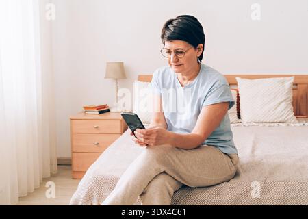 Mature caucasian woman using smartphone in cozy bedroom setting Stock Photo