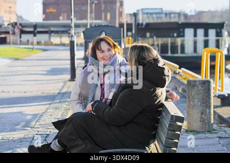 Women with brown and dark hair are seated on a bench in a sunny park, sharing a joyful conversation, surrounded by greenery and urban scenery Stock Photo