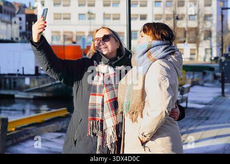 Two women are happily taking a selfie outdoors by the water, dressed warmly for the weather, showcasing a moment of friendship and shared joy in a vib Stock Photo