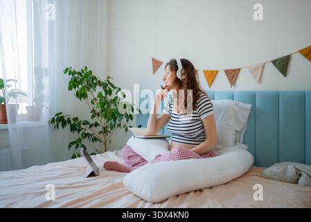 Pregnant woman comfortably sitting on her bed, wearing headphones and using a tablet to participate in an online lecture about childbirth, diligently taking notes in a notebook for birth education Stock Photo