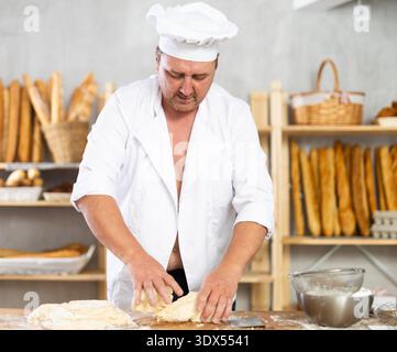Male baker in unbuttoned uniform prepares dough, kneads it with his hands Stock Photo