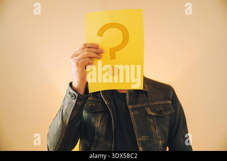 Man holding yellow question mark sign covering face, mysterious portrait selective focus Stock Photo