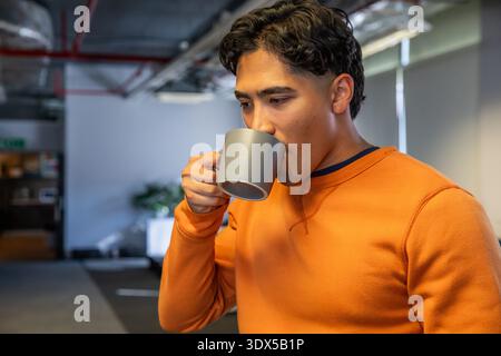 Asian man standing in open office sipping from grey mug while wearing orange sweatshirt, copy space Stock Photo