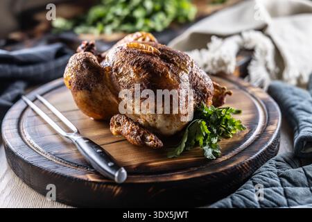 A whole chicken, baked until golden brown, is served on a wooden cutting board, garnished with fresh parsley and accompanied by a meat fork in a rusti Stock Photo