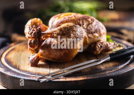 A whole chicken, baked until golden brown, is served on a wooden cutting board, garnished with fresh parsley and accompanied by a meat fork in a rusti Stock Photo