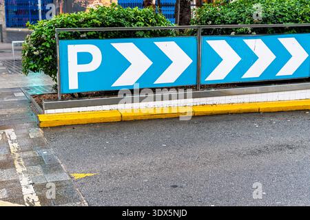 Blue parking direction sign with white p and arrow chevrons beside curb on urban street with greenery. Stock Photo