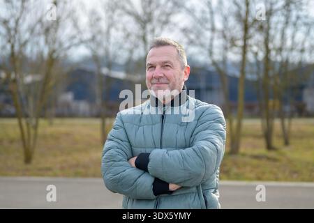 Mature man in a padded jacket standing outdoors with crossed arms, looking confident and relaxed in a natural park environment. Stock Photo