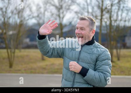 Smiling mature man with open mouth waving his hand outdoors in a park, greeting someone and enjoying a bright, relaxed moment. Stock Photo
