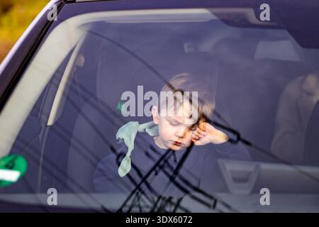 A tired toddler sleeping inside a car, seen through the windshield with soft reflections. The peaceful scene represents childhood exhaustion, travel, Stock Photo