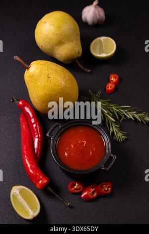 Pears, red chili peppers, garlic, rosemary and lime arranged around a small black bowl of sweet chili sauce on a dark tabletop, rustic overhead still Stock Photo