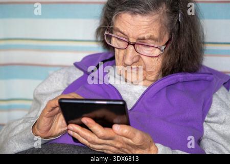 Elderly woman wearing glasses using tablet at home, focused on touchscreen and digital communication Stock Photo