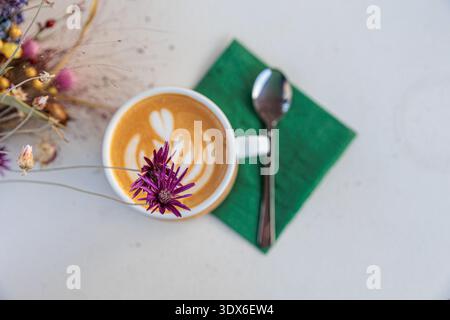 Warm latte with leaf latte art served in a white cup sits on a green napkin with a spoon, next to a colorful dried flower bouquet on a light tabletop, Stock Photo
