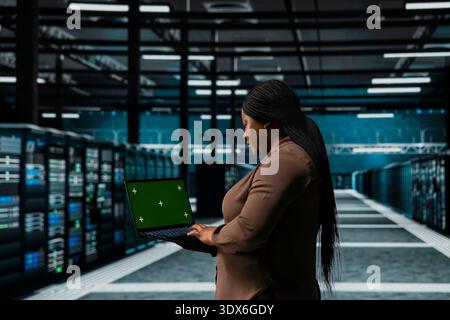 Woman uses green screen laptop in high tech data center to troubleshoot hardware. Worker uses chroma key device in server hub, running software development tools, resolving system crashes Stock Photo