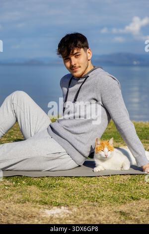 A young man in casual gray attire sits relaxed on a mat by a peaceful lakeside, sharing a tranquil moment with a white and orange cat on a sunny day. Stock Photo
