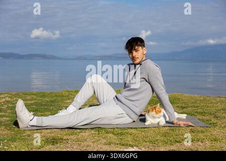 A young man in casual wear relaxes on a mat by a tranquil lake with a cat beside him. The serene landscape with clear skies and distant mountains adds Stock Photo