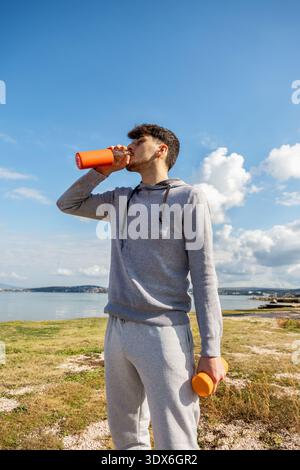 A young man in a grey tracksuit drinks water from an orange bottle, standing outdoors with a scenic sea view. The sky is clear, and the atmosphere is Stock Photo