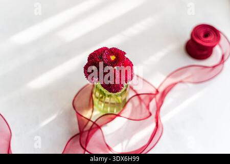 A bright spring scene featuring vibrant red flowers in a vase, accompanied by a delicate red ribbon elegantly spread across a white table, creating a Stock Photo