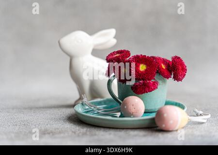 A festive Easter table setting features a ceramic bunny, a turquoise cup filled with vibrant red flowers, and decorative pink eggs, all evoking the sp Stock Photo
