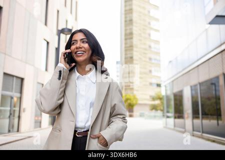 A cheerful Hispanic Colombian businesswoman in a beige blazer smiles while talking on her smartphone, walking confidently through an urban business di Stock Photo