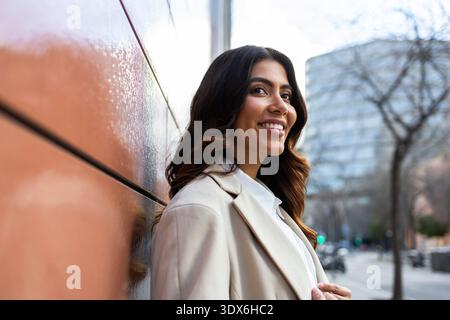 Hispanic businesswoman standing confidently outside against a modern building wall. She embodies professionalism and success, wearing a beige blazer a Stock Photo