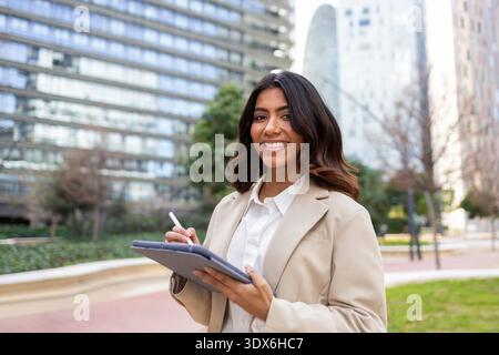 Hispanic businesswoman wearing a beige blazer, smiling while holding a tablet and stylus. She stands outdoors in a modern office park with skyscrapers Stock Photo
