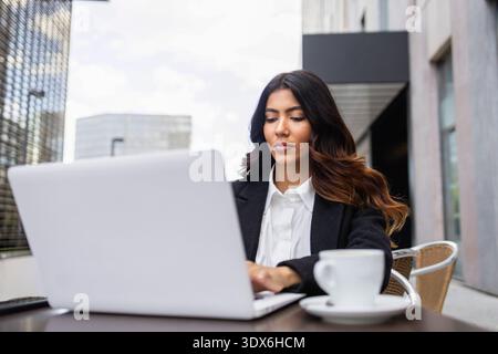 Hispanic businesswoman sitting at an outdoor table, intensely focused on her laptop screen. Colombian woman, dressed in business attire, with a cup of Stock Photo