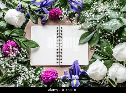 Flatlay image of an open spiral notebook surrounded by vibrant purple and blue flowers, lush green leaves, and delicate white blooms, creating a fresh Stock Photo