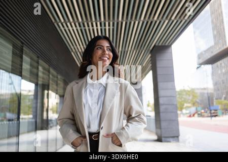 A Hispanic businesswoman walking confidently through a modern urban environment, wearing a smart beige blazer. The cityscape reflects her professional Stock Photo