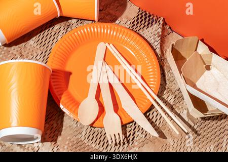 A vibrant set of disposable tableware featuring an orange paper plate, matching paper cups, and wooden utensils. Ideal for eco-friendly gatherings, pi Stock Photo