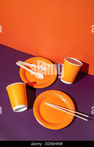 Bright orange disposable tableware set featuring paper plates, cups, and wooden utensils on a contrasting purple background, ideal for vibrant, eco-fr Stock Photo
