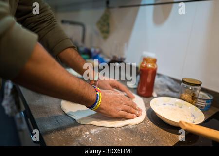A man is preparing homemade pizza in his kitchen. His hands are seen working the dough on a wooden countertop with ingredients like sauce and seasonin Stock Photo