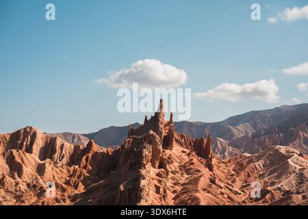 Tall red rock spire rising from Fairy Tale canyon in Kyrgyzstan. Dramatic desert landscape with mountain backdrop and scattered cloud in bright daylig Stock Photo