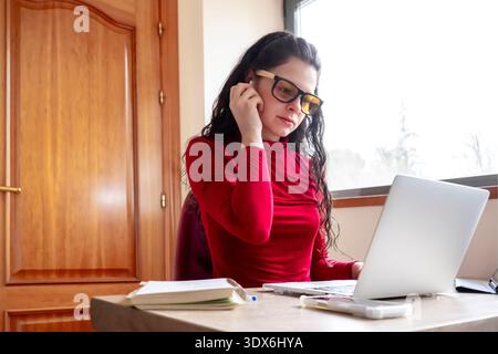 A focused woman wearing glasses works at a laptop in a bright office. She is seated at a desk with scattered notebooks, deeply engaged in her tasks, r Stock Photo