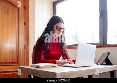 A woman wearing glasses works focused on her laptop, seated at a desk in a bright office. The sunlight streams through a large window, creating a warm Stock Photo