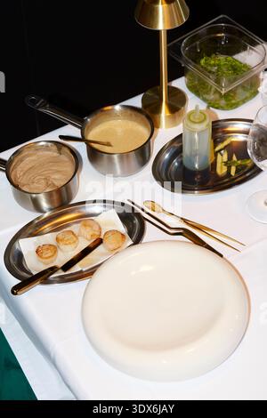 A gourmet restaurant setting with a white tablecloth showcasing seared scallops, various sauces in steel pans, garnishes, and elegant gold utensils, r Stock Photo
