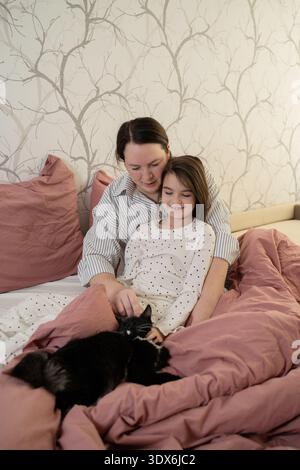 A mother and her daughter enjoy a cozy moment together on the bed, surrounded by pillows and blankets, while petting a cat. The warm home setting emph Stock Photo