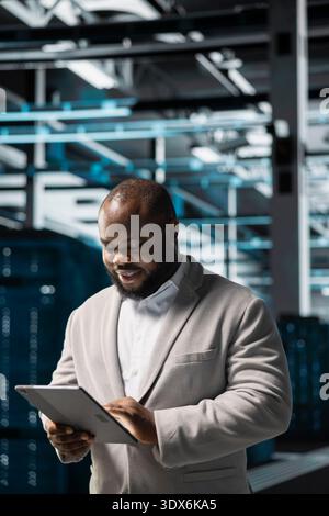 Cheerful technician in data center workspace running system diagnostic tests on tablet to fix problems. Joyous employee looking for network issues affecting rigs performance using device Stock Photo